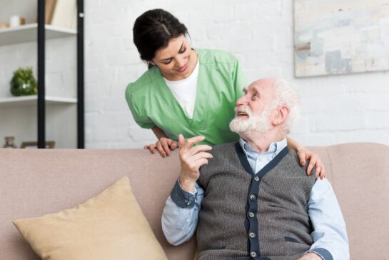 Male senior sitting on a couch smiling while looking at female nurse