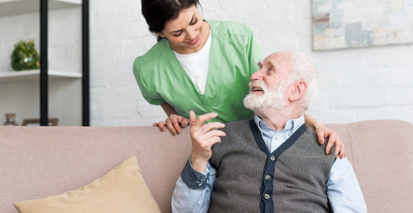 Male senior sitting on a couch smiling while looking at female nurse