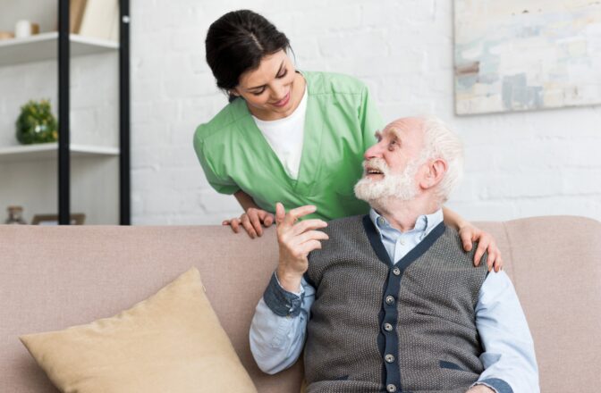 Male senior sitting on a couch smiling while looking at female nurse