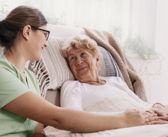 Female healthcare professional sitting next to elder woman while both of them hold hands and smile