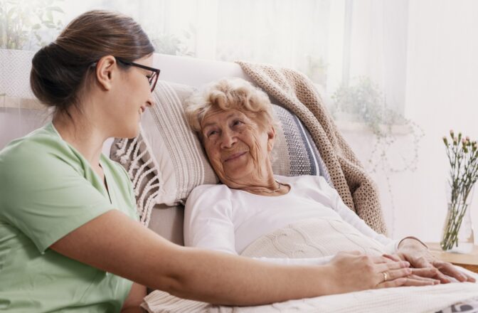 Female healthcare professional sitting next to elder woman while both of them hold hands and smile