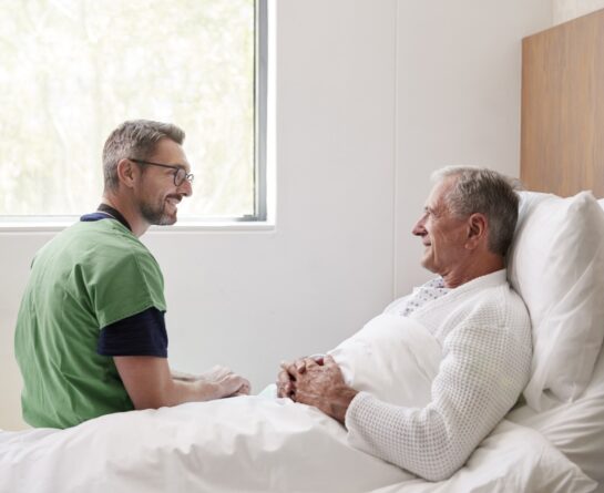 Male healthcare professional sitting next to elder man while both of them look at each other and smile