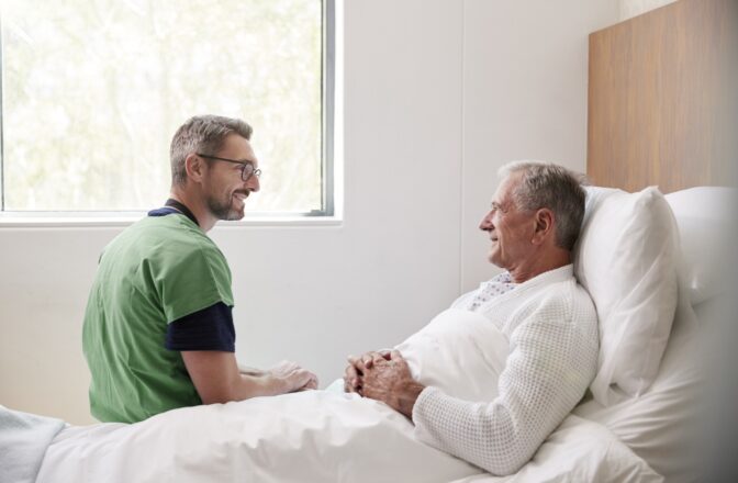 Male healthcare professional sitting next to elder man while both of them look at each other and smile