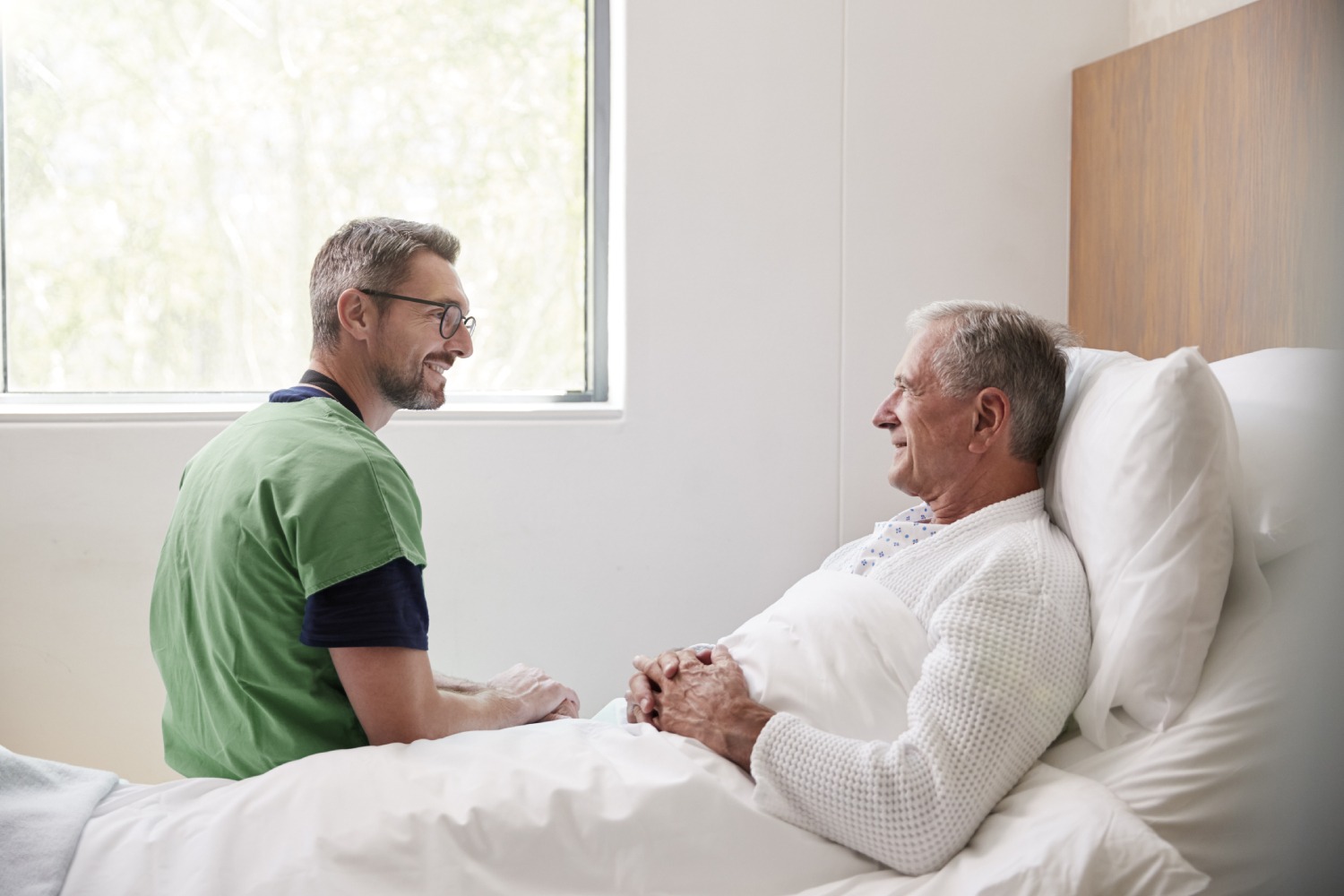 Male healthcare professional sitting next to elder man while both of them look at each other and smile