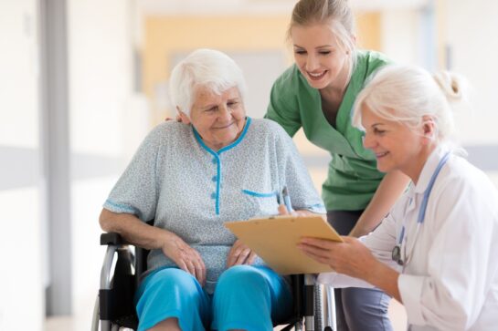 mature woman in wheelchair being assisted by a healthcare professional and listening to a second healthcare professional with clipboard and stethoscope