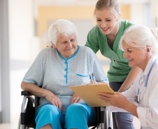 mature woman in wheelchair being assisted by a healthcare professional and listening to a second healthcare professional with clipboard and stethoscope