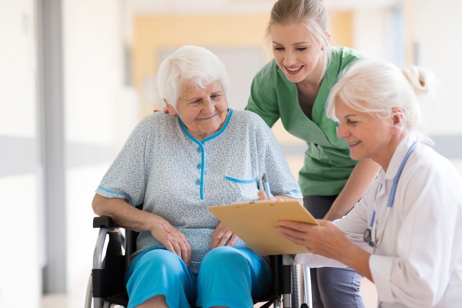 mature woman in wheelchair being assisted by a healthcare professional and listening to a second healthcare professional with clipboard and stethoscope