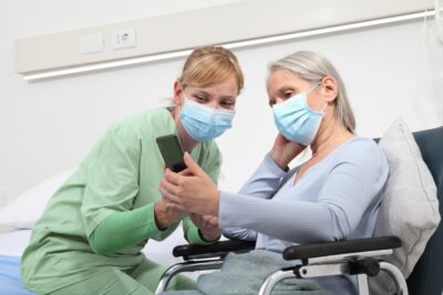 Mature woman in wheelchair wearing face mask looking at cell phone with a healthcare professional wearing a face mask