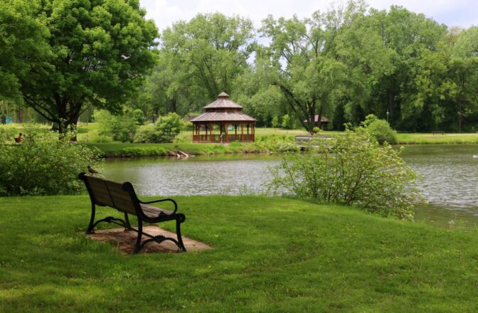 wood and metal bench in green surroundings near stream with gazebo across water