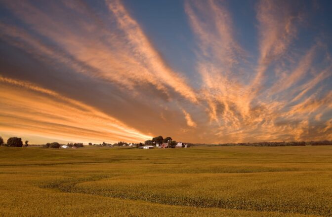 open field with blue cloudy sky and farm in the distance