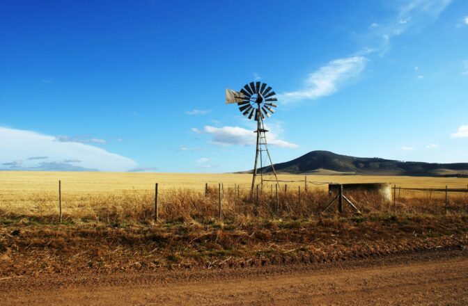 windmill near wire fence along dirt road with bluffs in the distance