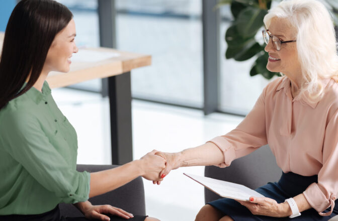 Elder woman wearing glasses handshakes younger woman as they're sitting face to face