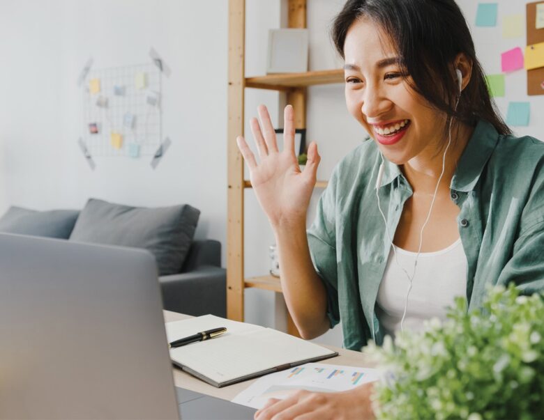 Woman smiles and waves at laptop while wearing headphones