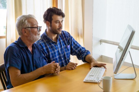 Male senior sits next to younger male as they both look into the computer's screen