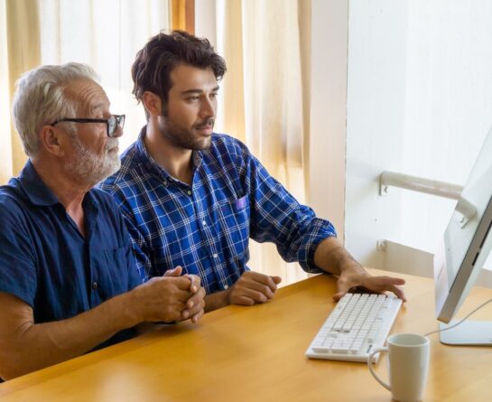 Male senior sits next to younger male as they both look into the computer's screen
