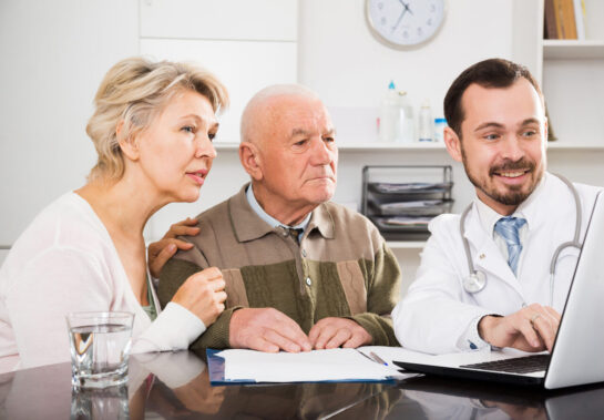Elder couple and doctor sit on a table looking at laptop's screen