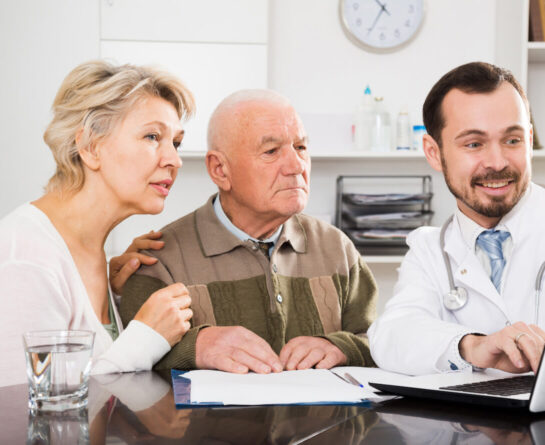 Elder couple and doctor sit on a table looking at laptop's screen