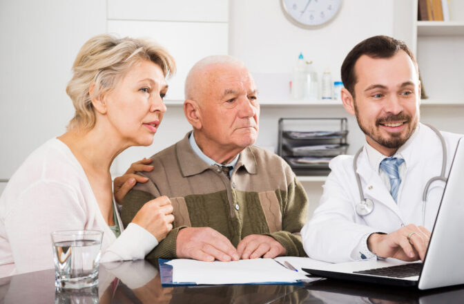 Elder couple and doctor sit on a table looking at laptop's screen