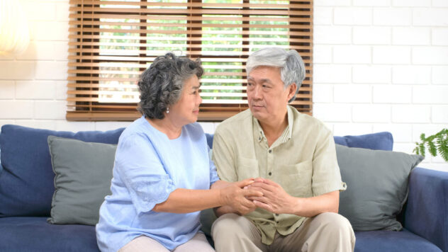 Elder couple sit next to each other while holding hands on blue couch