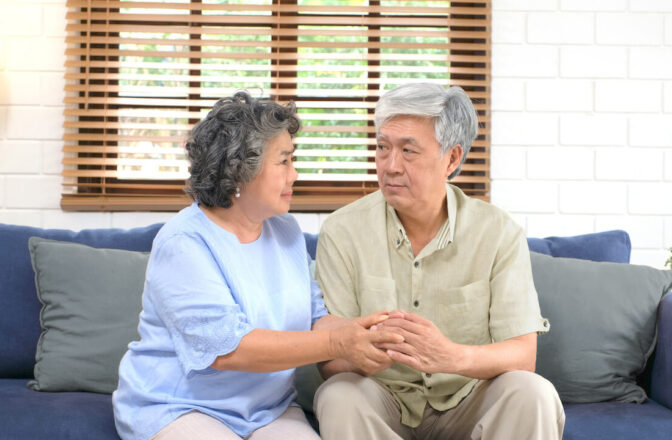 Elder couple sit next to each other while holding hands on blue couch