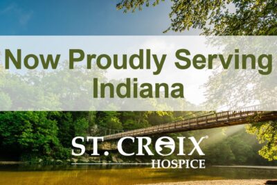 Text informing that St. Croix Hospice now serves Indiana area with wooden bridge crossing a lake as background
