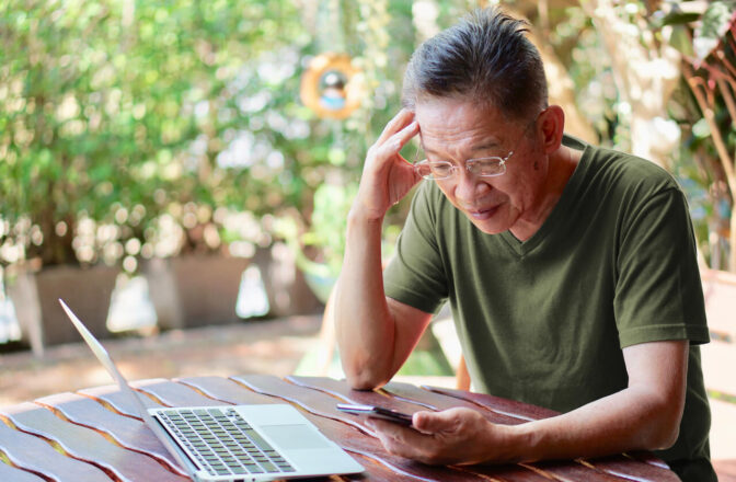 Senior man checking his phone while sitting on a table with laptop open