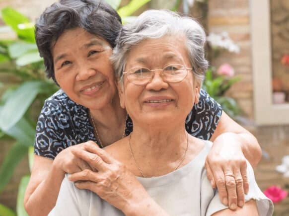 Two Asian elder women smiling while one holds the other one's hand in a friendly way