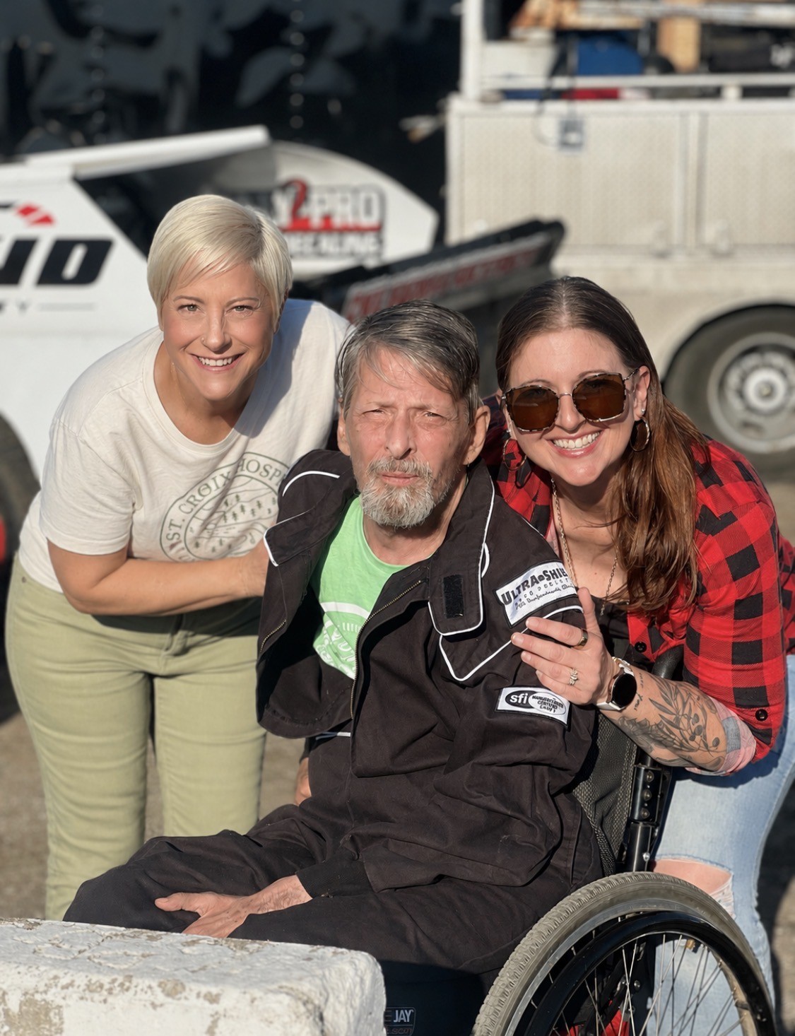man wearing racing jacket in wheelchair surrounded by two supporters smiling
