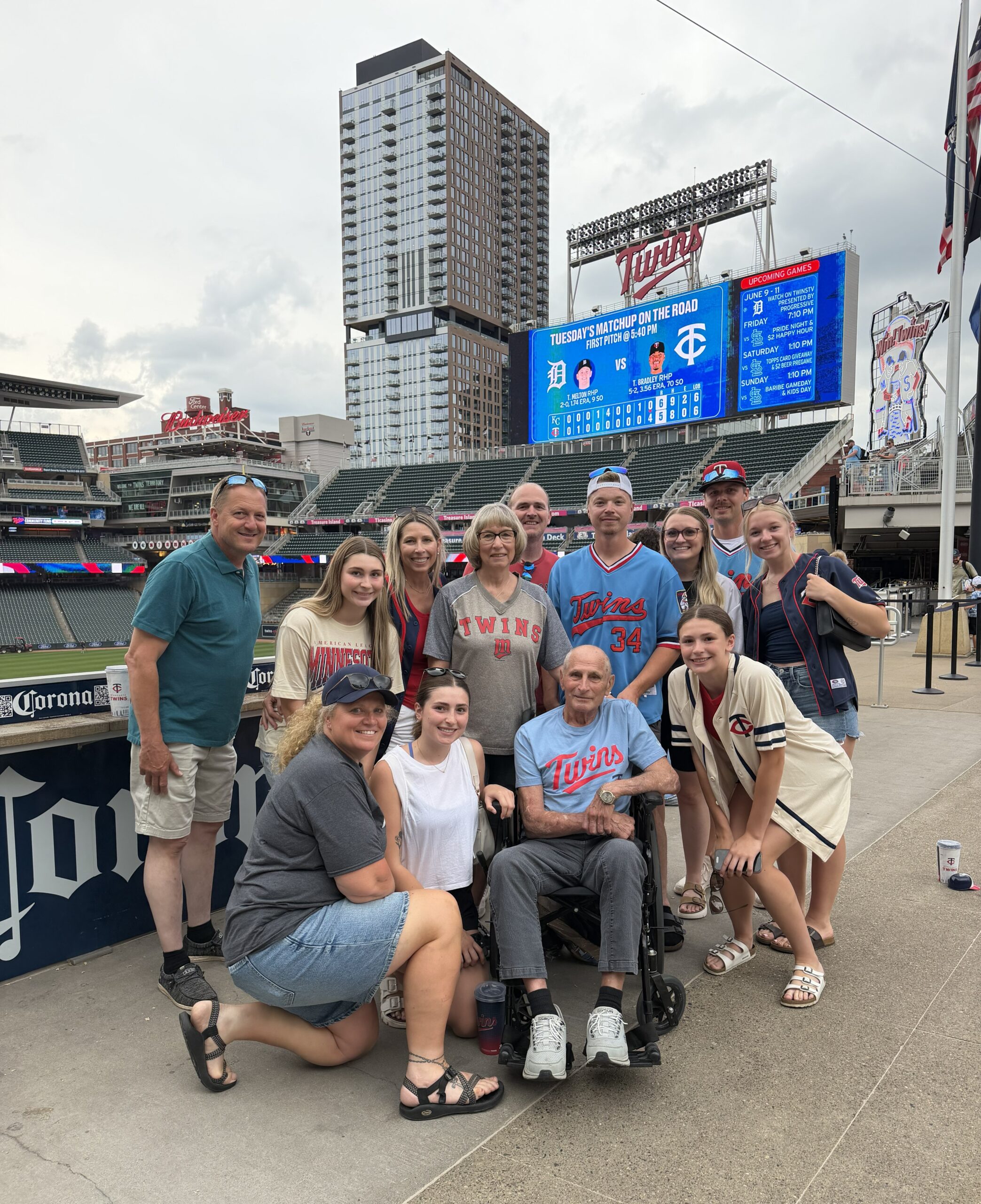 Group of family members in Twins gear gather around man in wheelchair on stadium plaza with scoreboard and stadium in background