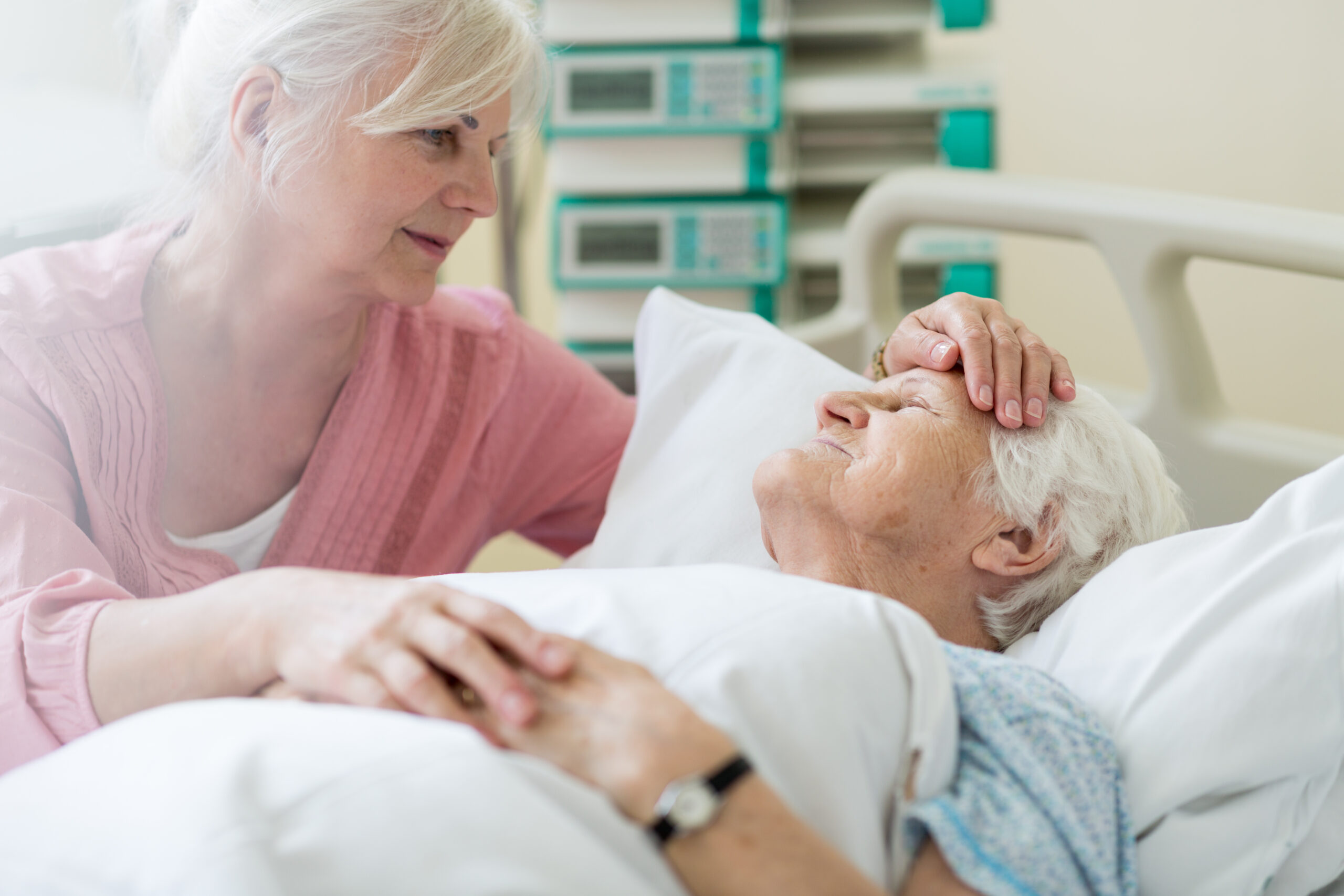 woman comforting elderly patient in bed holding her hand and caressing forehead